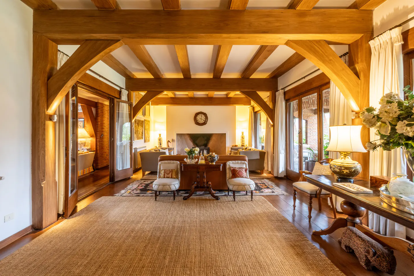 A living room with exposed wooden beams, a fireplace, and neutral-toned furniture. Natural light streams in through open glass doors.