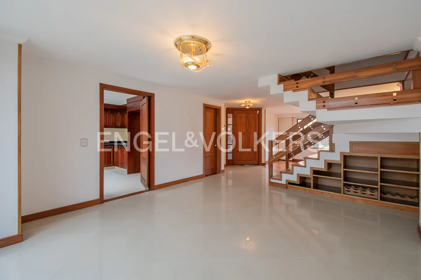 Bright, open-plan living area with white tile floors, wood trim, and staircase with built-in shelving. A doorway leads to a kitchen.