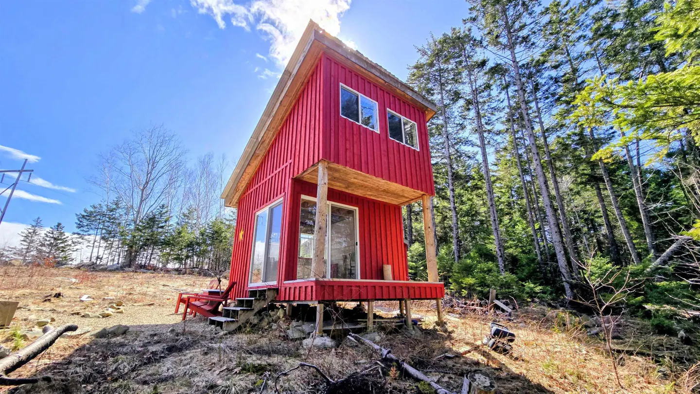 A small, two-story red cabin with a porch sits on a hill surrounded by trees under a sunny blue sky.