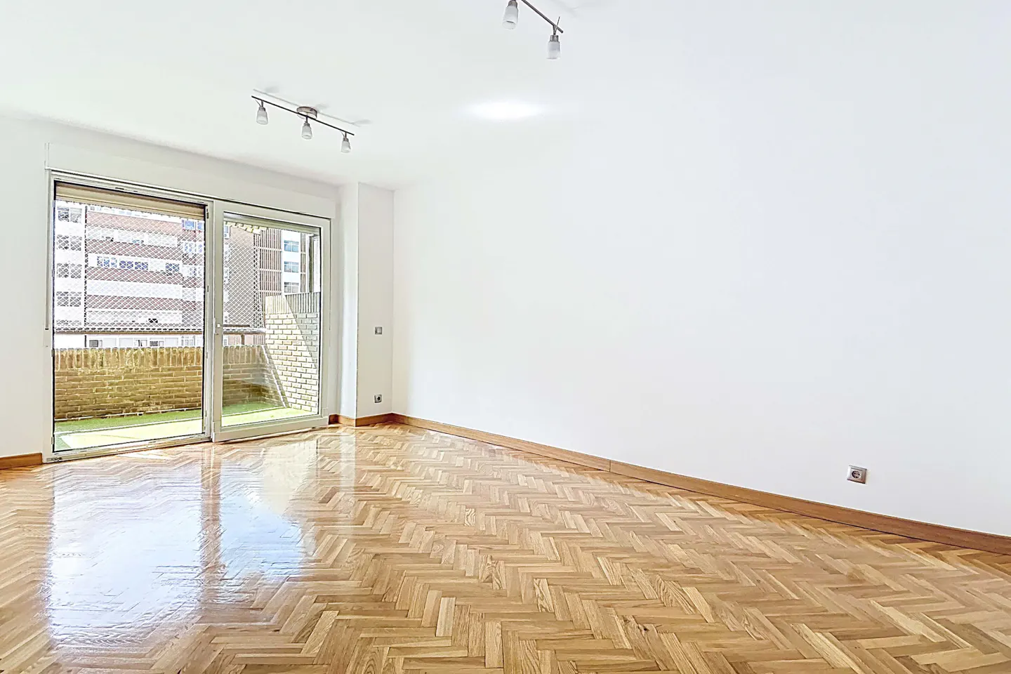 Bright, empty room with herringbone wood floor, white walls, and sliding glass doors to a balcony with brick and greenery.