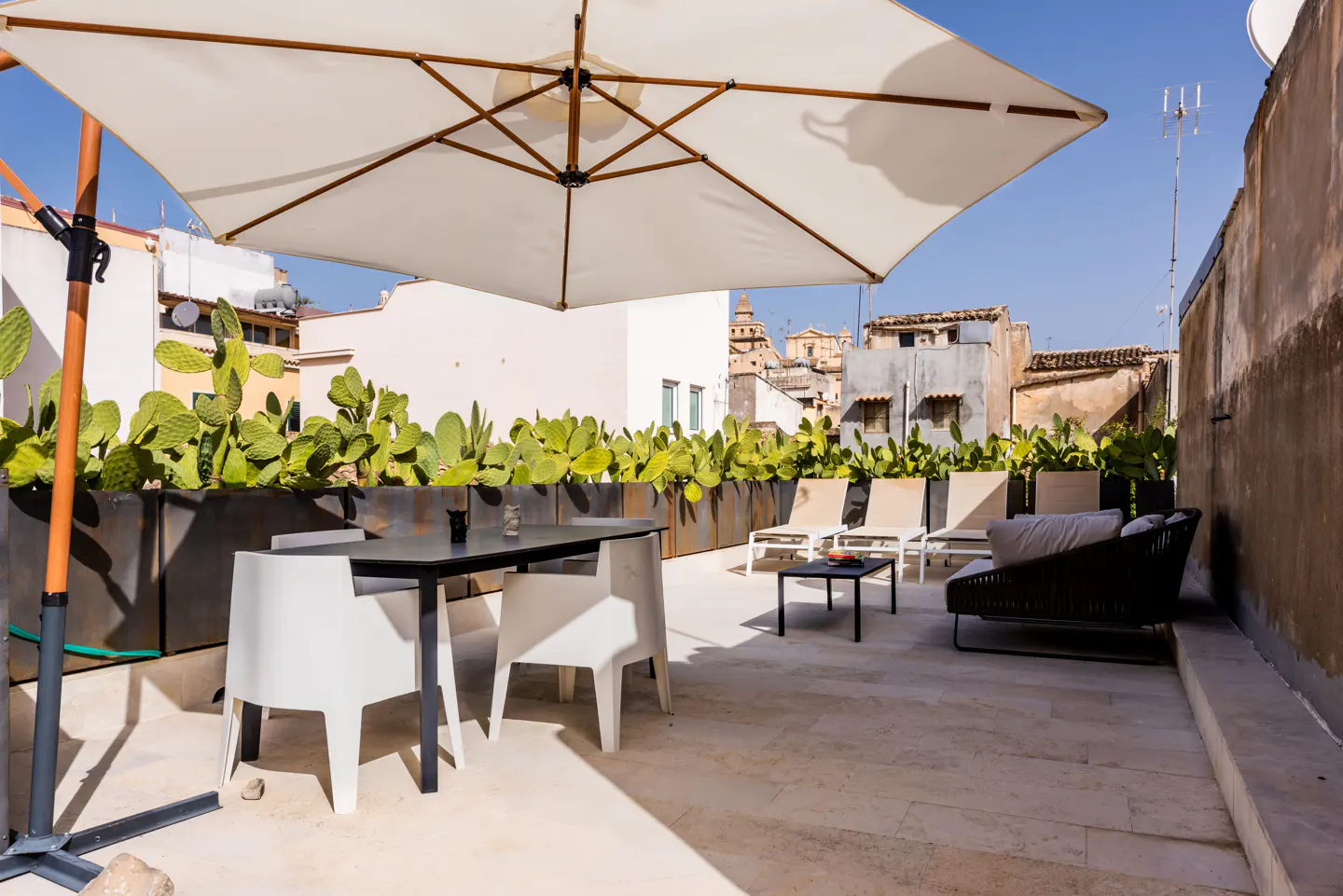 Rooftop patio with a large white umbrella, table, chairs, lounge chairs, and cactus planters. Buildings are visible in the background.