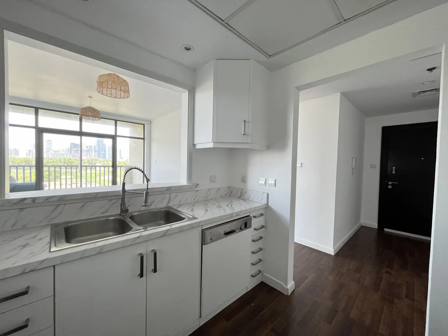A bright kitchen with white cabinets, marble countertops, and a stainless steel sink. A large window overlooks a green park and city skyline. Dark wood floors lead to a black door.