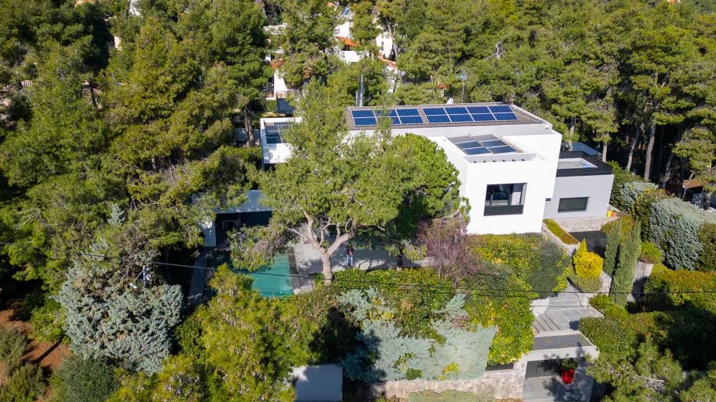 Aerial view of a modern white house with solar panels, surrounded by lush green trees and landscaping.