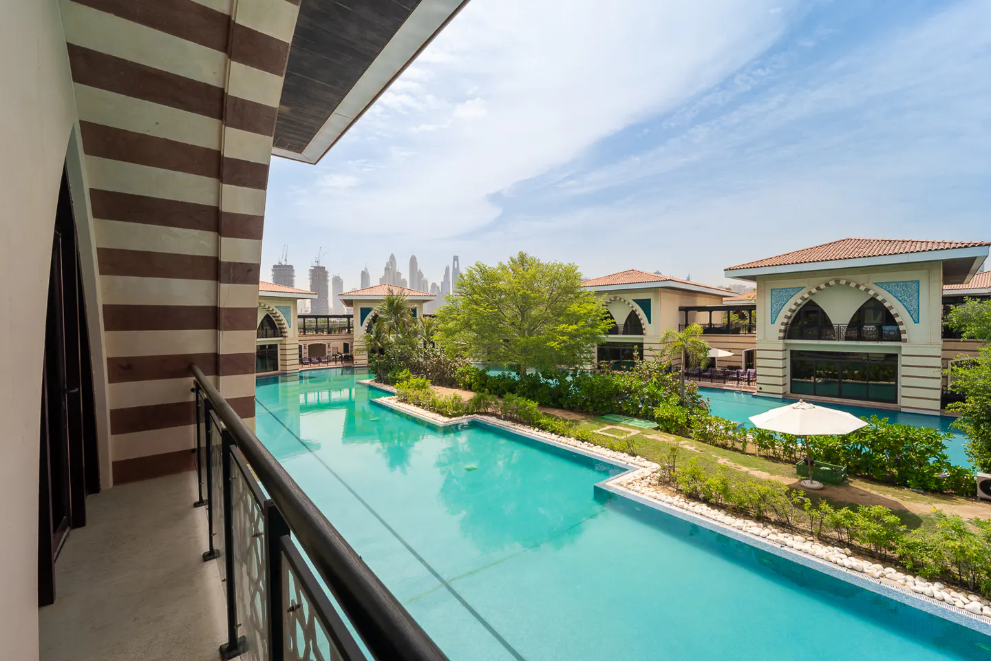 View from a balcony overlooking a turquoise pool, lush greenery, and buildings with arched windows. Distant city skyline under a blue sky.