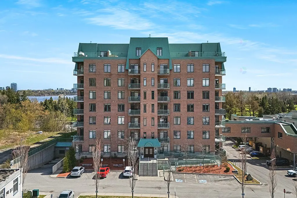 Exterior view of a brick apartment building with a green roof and balconies, cars parked in front.