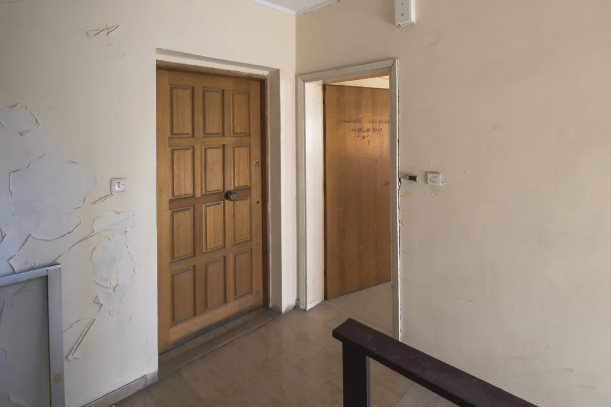 Interior view of a hallway with a brown wooden door and an open doorway. The walls are peeling and off-white.