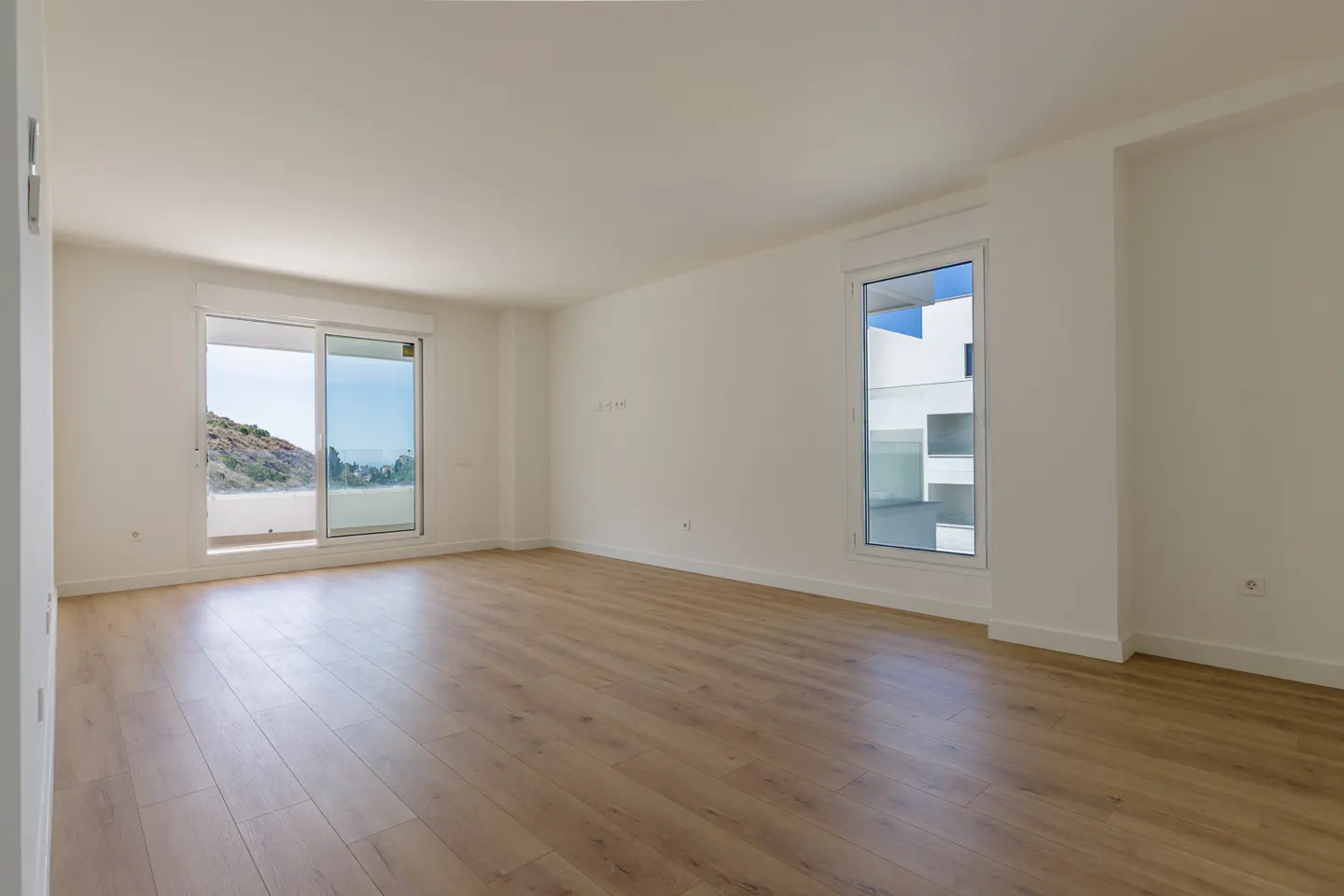 Bright, empty room with light wood floors, white walls, and two windows. One window is a sliding glass door with a view of a hill and ocean.