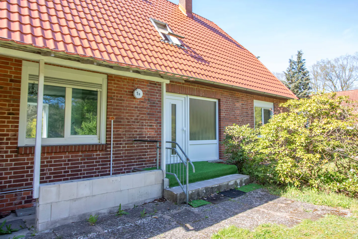 Exterior view of a one-story brick house with a red tile roof, white door, and green lawn.