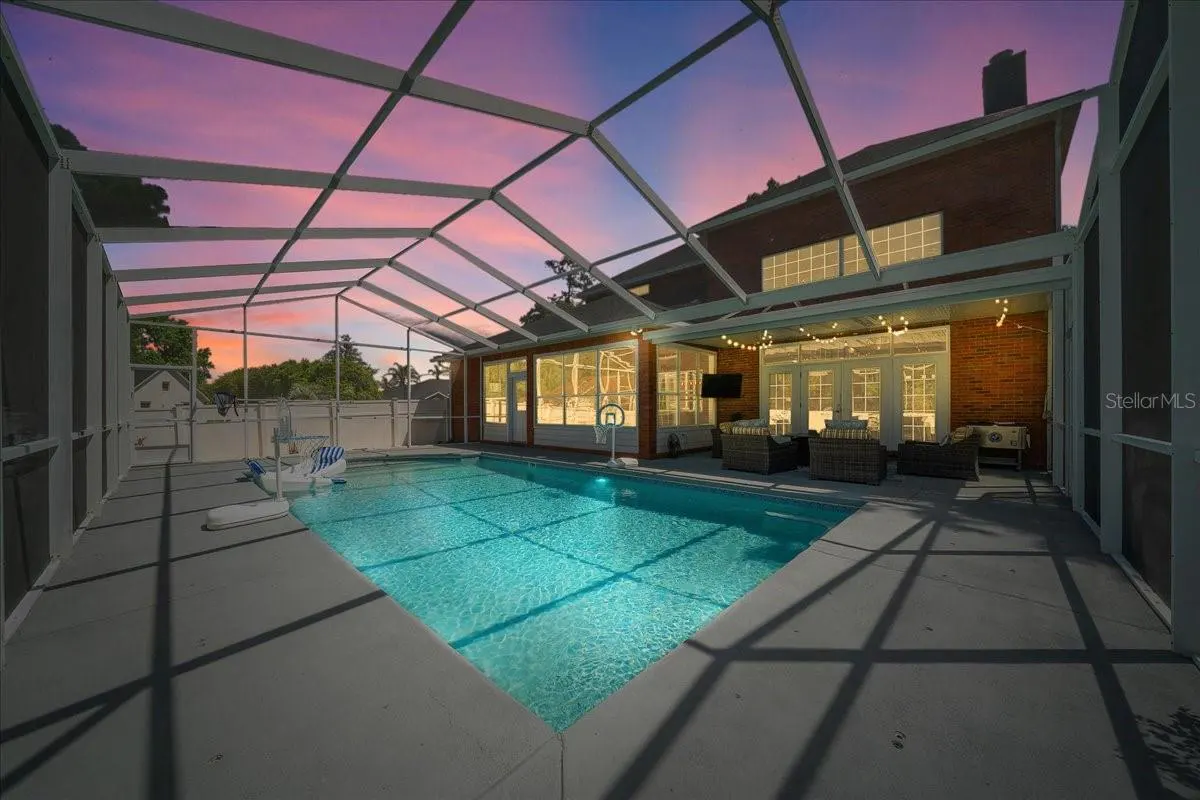 Screened pool with turquoise water, float, and basketball hoop. Brick house with patio furniture and string lights in the background. Dusk sky.