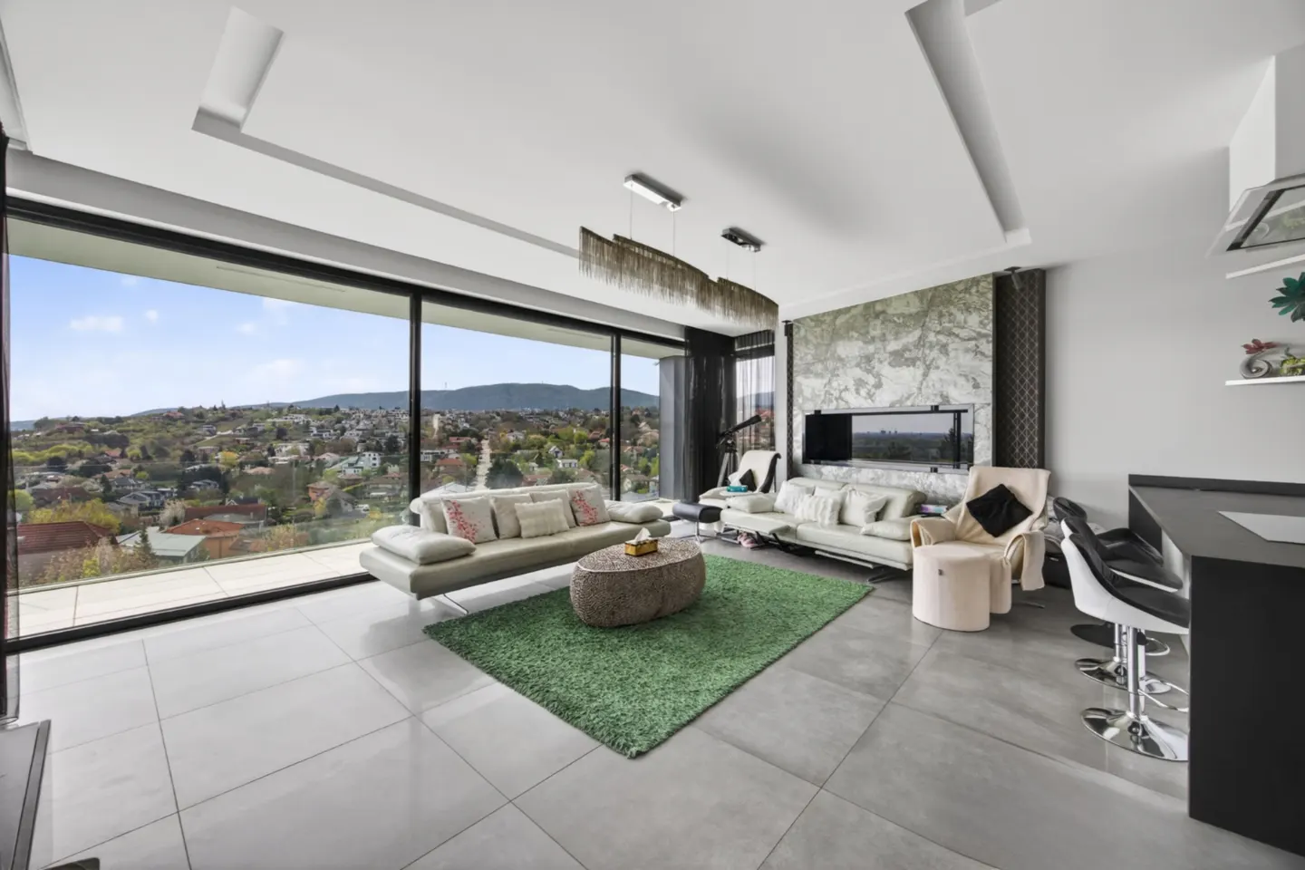 Modern living room with a large window view. Two sofas face each other on a green rug. A black bar with stools is on the right.
