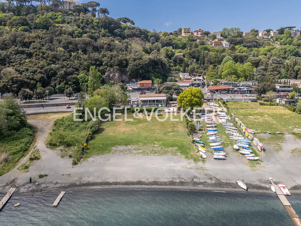 Aerial view of a lakefront property with boats, a beach, and a grassy area, backed by a tree-covered hillside and buildings.