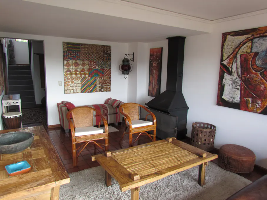Living room with rattan chairs, bamboo table, and black fireplace. Colorful art adorns the white walls. A staircase is visible in the background.