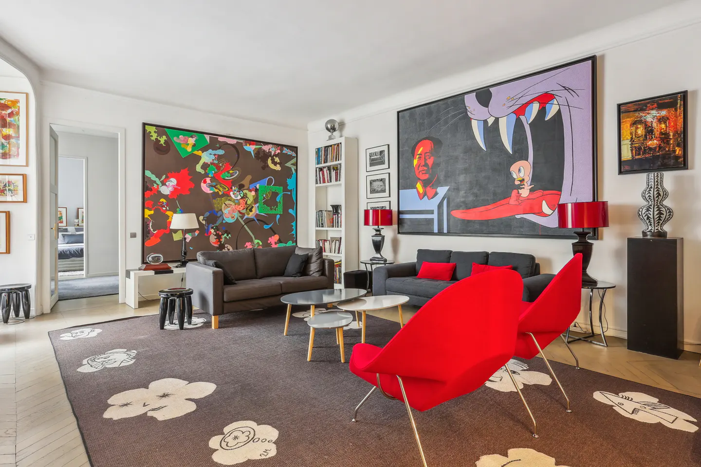 Living room with two gray sofas, red chairs, and large abstract art. A brown rug with white flower designs covers the floor.