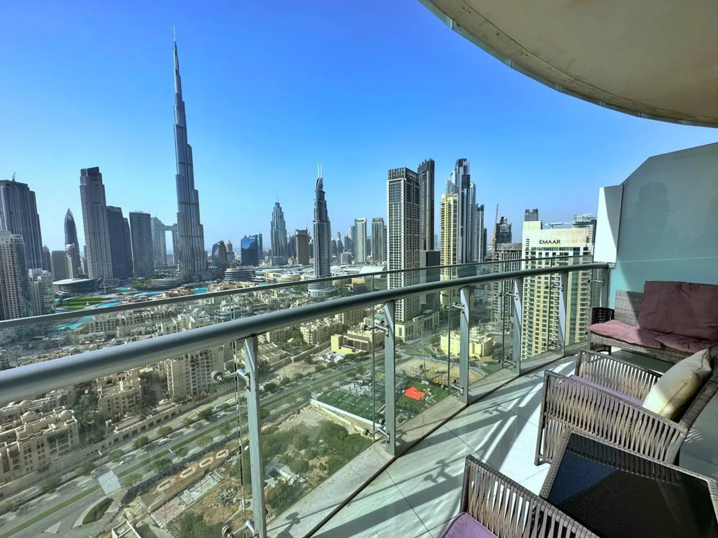 Balcony view of Dubai skyline. Wicker chairs with purple cushions face the city. Burj Khalifa is visible against a clear blue sky.