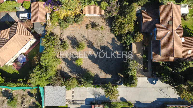 Aerial view of a vacant lot surrounded by houses with red tile roofs and green trees. The Engel & Völkers logo is superimposed on the lot.