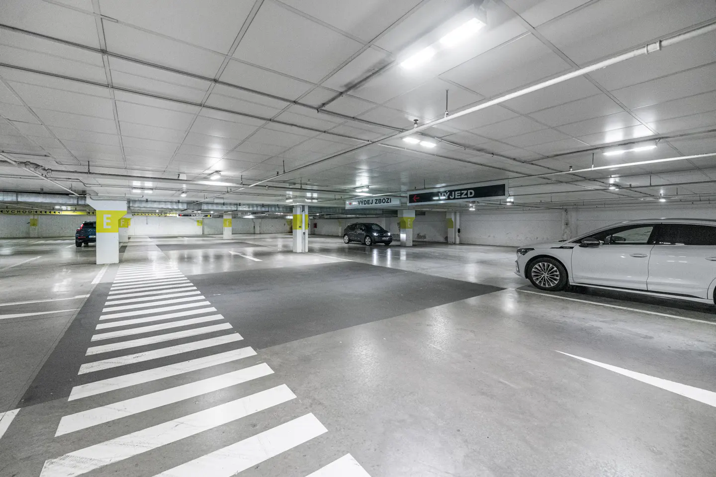 Indoor parking garage with white ceiling, gray floor, and white striped crosswalk. Cars are parked in the background and foreground.