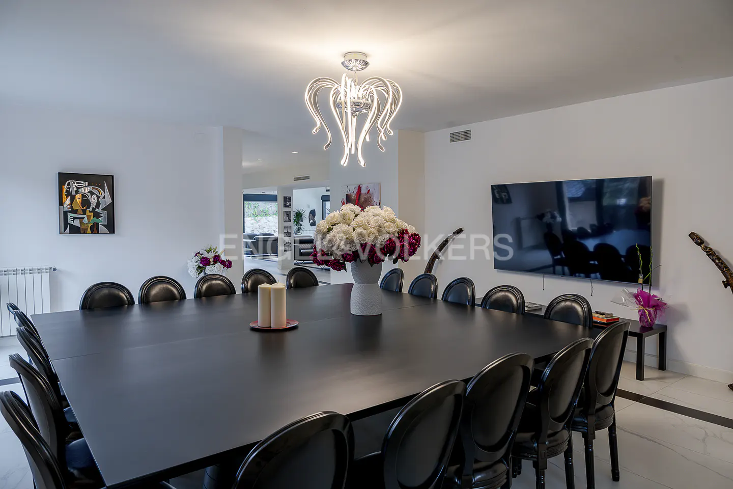 A modern dining room with a long black table, black chairs, a white flower arrangement, and a unique chandelier.
