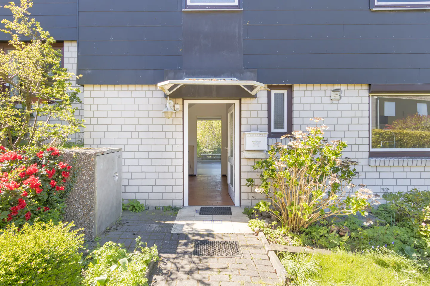 Exterior of a two-story house with white brick and dark siding, an open front door, and a brick walkway.