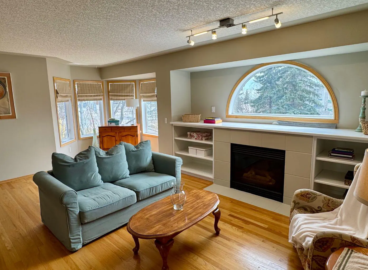 Living room with hardwood floors, a blue sofa, and a fireplace with built-in shelves. A large arched window overlooks trees.