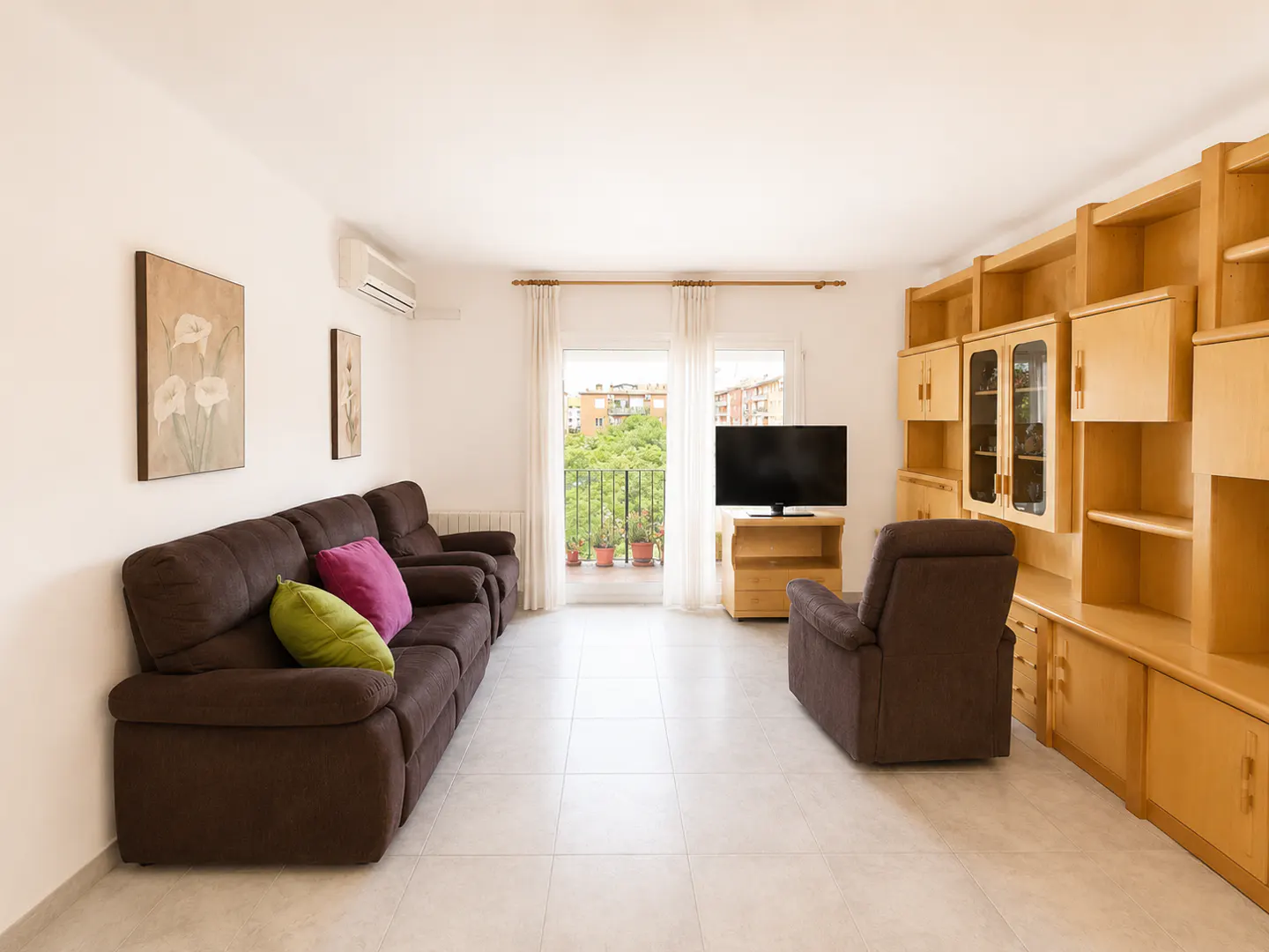 Bright living room with brown sofa, chair, and wood shelving. Balcony doors open to a view of trees and buildings.