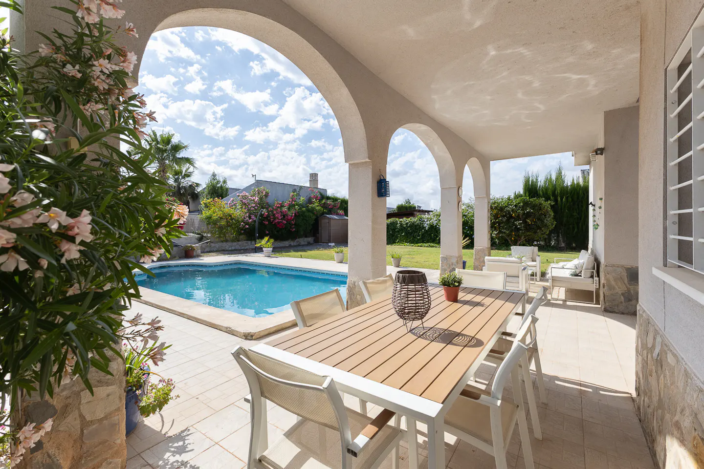 Covered patio with dining table and chairs overlooking a sunny backyard swimming pool and lush garden.