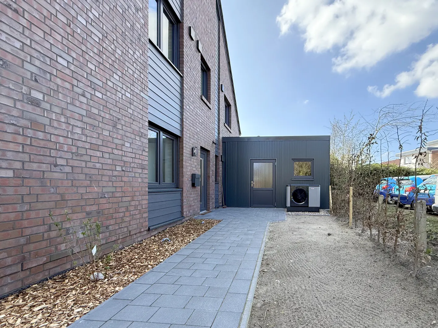 Exterior view of a brick house with a gray walkway leading to a gray outbuilding with a door and a heat pump.
