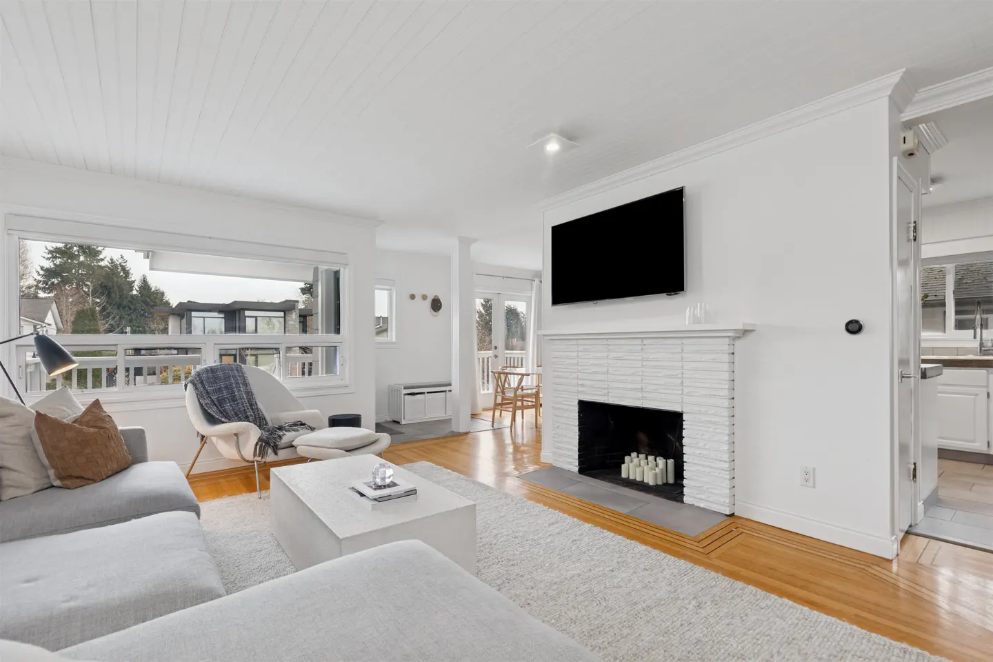 Bright living room with white walls, wood floors, and a large window. A gray sectional sofa faces a white brick fireplace with a mounted TV.