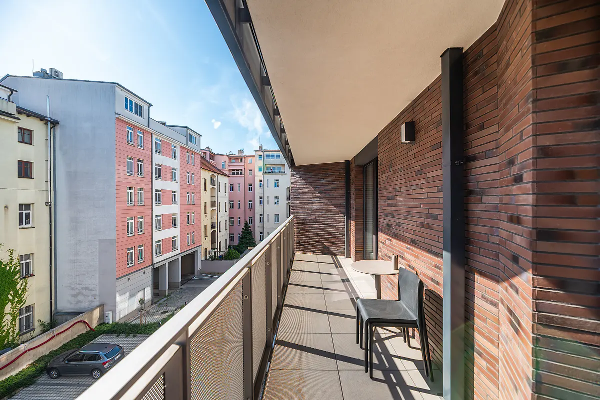 Modern balcony with dark brick walls, a small table, and chairs, overlooking a city street with colorful apartment buildings.