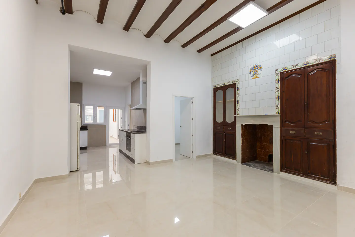 Bright, open-concept living space with white walls, tile floors, and dark wood ceiling beams. A kitchen and fireplace are visible.