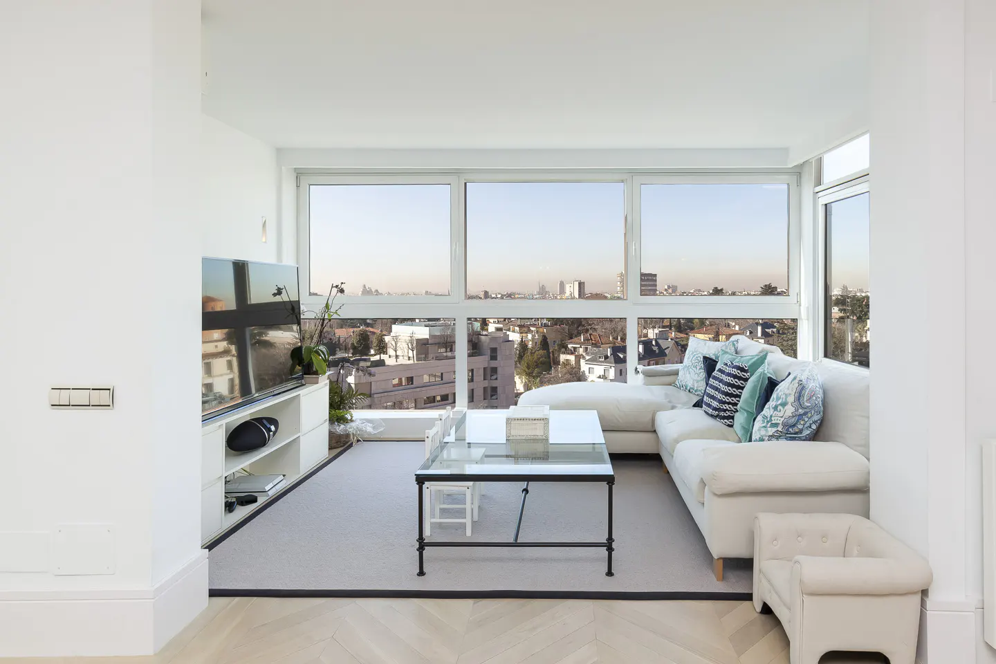 Bright living room with a white sectional sofa, glass coffee table, and large windows overlooking a city.