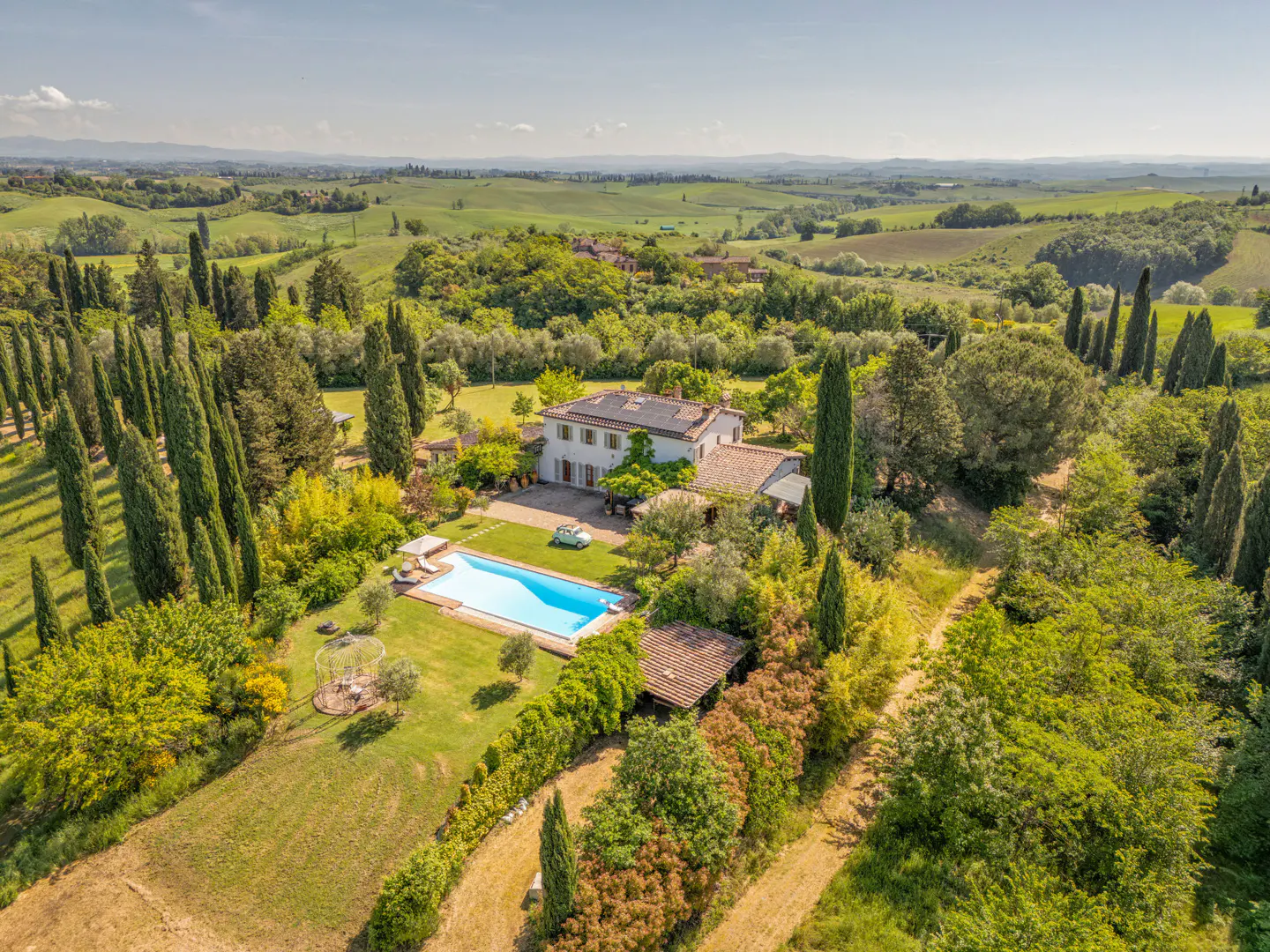 Aerial view of a Tuscan villa with a pool, surrounded by green trees and rolling hills under a blue sky.