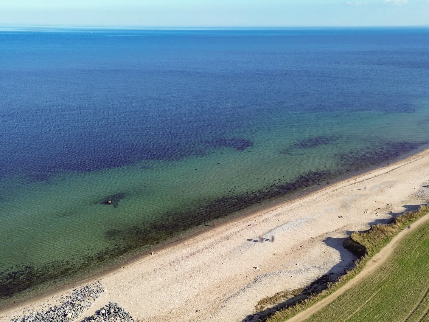 Aerial view of a sandy beach meeting a blue ocean, with green fields on the right.