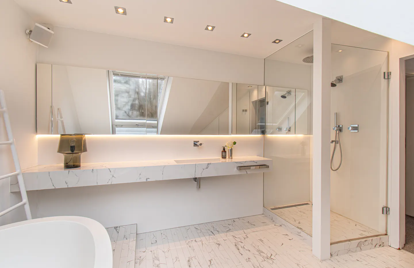 Bright, modern bathroom with marble floors, a long vanity with a mirror, and a glass-enclosed shower. A white ladder leans against the wall.
