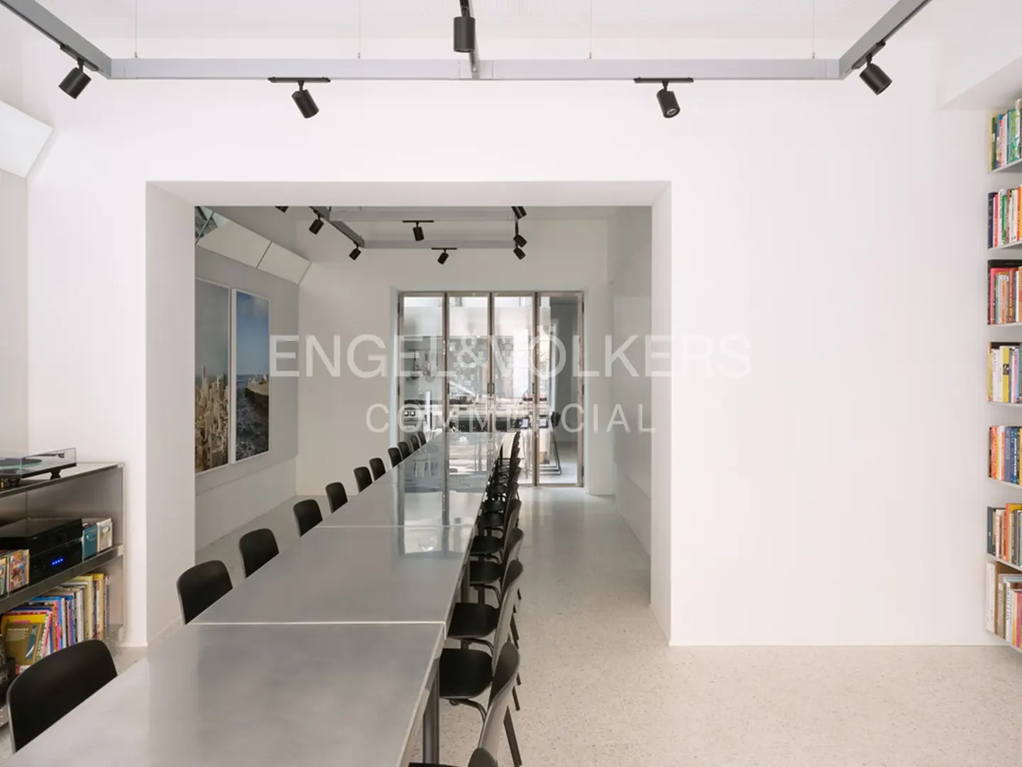 Bright, modern office space with a long table and black chairs. Bookshelves line the right wall. Track lighting illuminates the room.