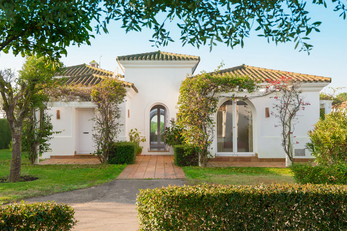 White house with arched windows and a red tile roof, surrounded by green grass and bushes.