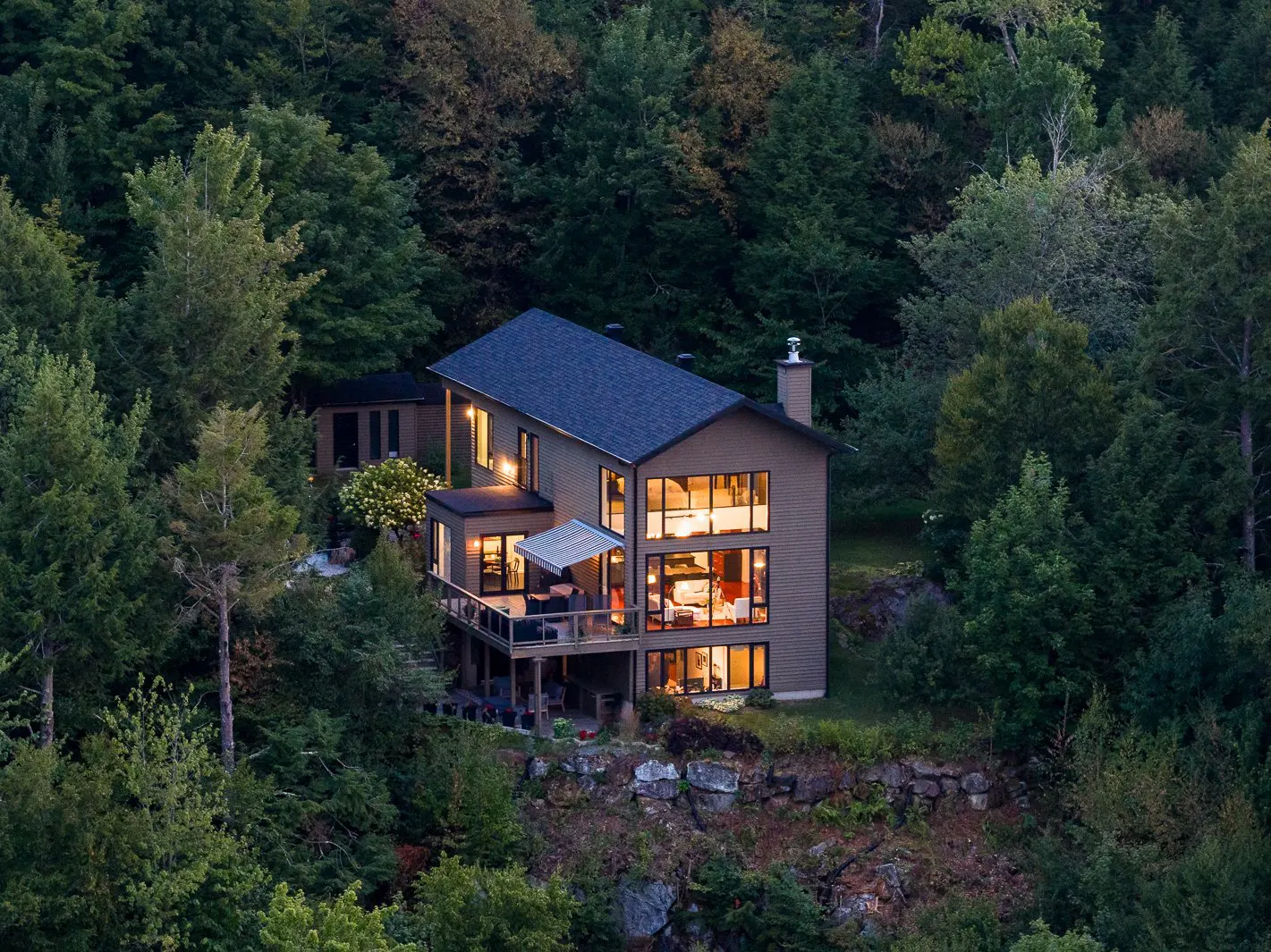 A modern, three-story house with large windows sits nestled in a dense green forest.