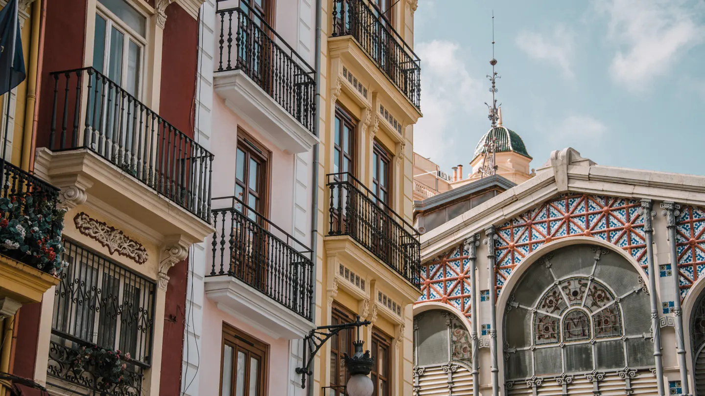 A wide street view with classic residential buildings on both sides and a large domed church with multiple towers in the background. Green trees partially obscure the building, and the sky is bright with light clouds.