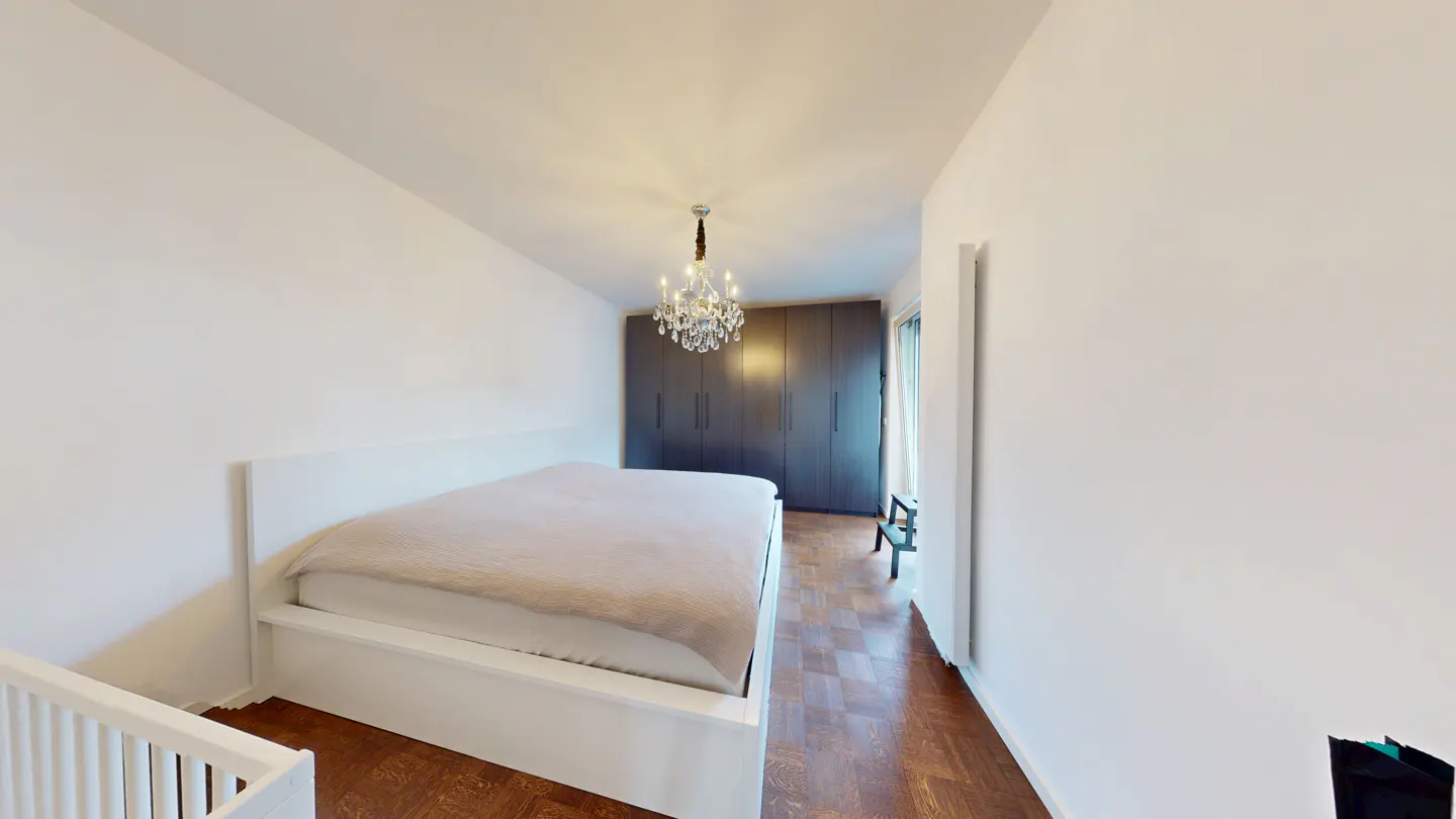 Bright bedroom with a white bed, wood floors, and a crystal chandelier. A gray wardrobe stands against the back wall.