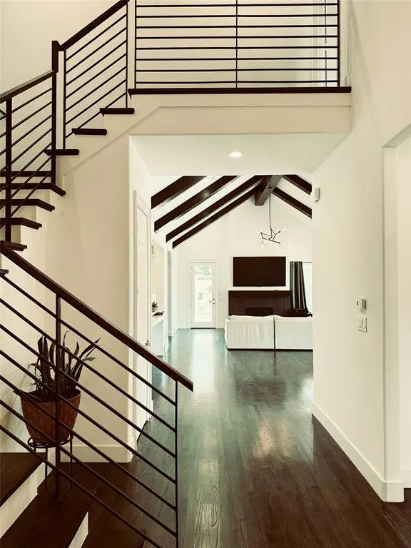 Interior view of a home's entry with dark wood floors, white walls, and a staircase with black metal railings. A living room is visible with a white sofa and vaulted ceiling.