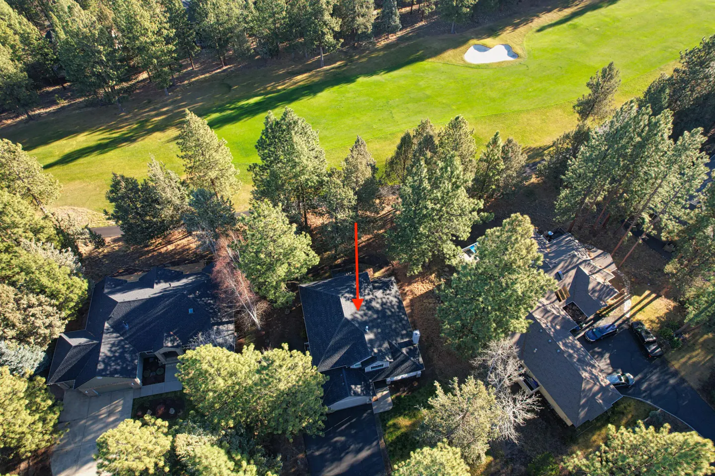 Aerial view of a dark-roofed house surrounded by green trees, next to a golf course with a white sand trap.