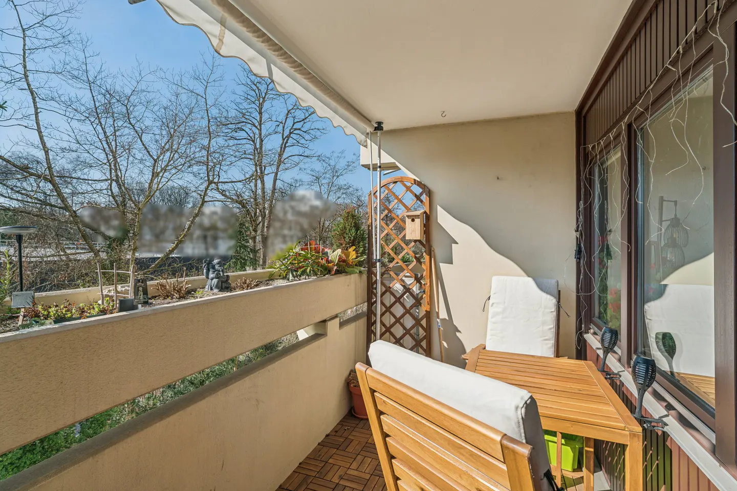 A sunny balcony with wood furniture, a trellis, and a planter box filled with greenery. Trees are visible in the background.