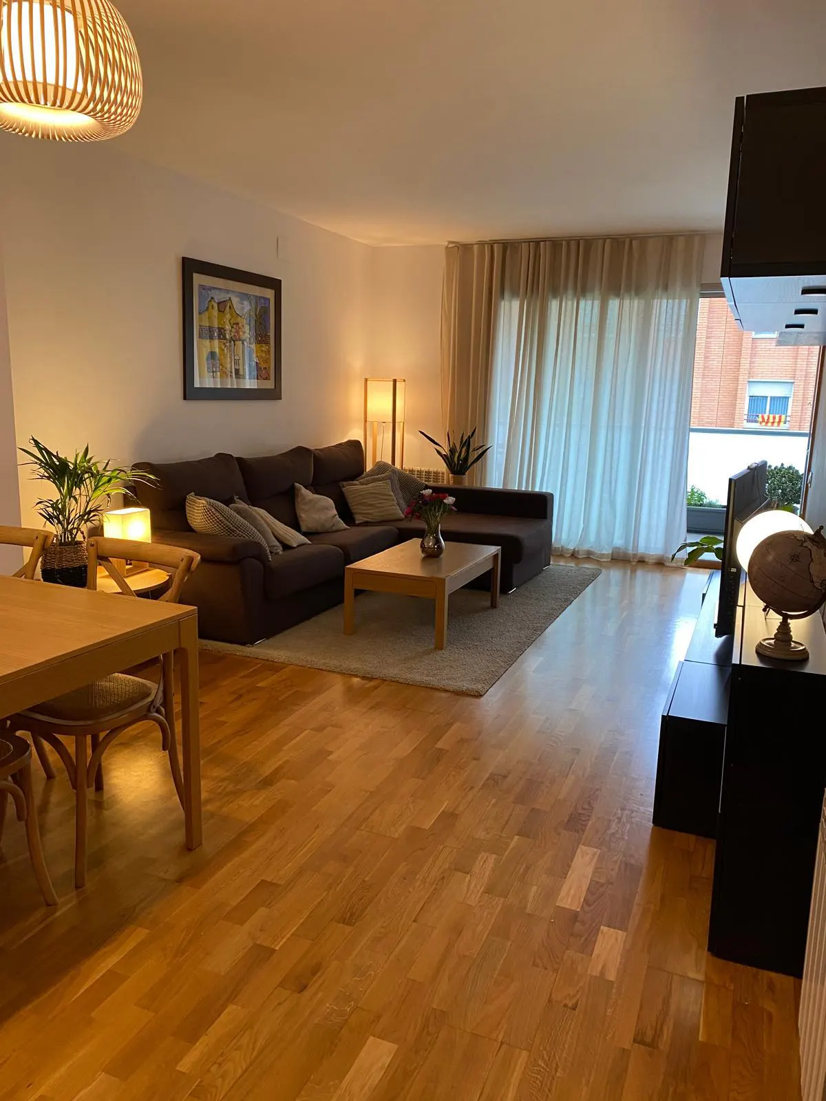 Living room with hardwood floors, a brown sectional sofa, a wooden coffee table, and a dining table with chairs. Curtains cover a window.