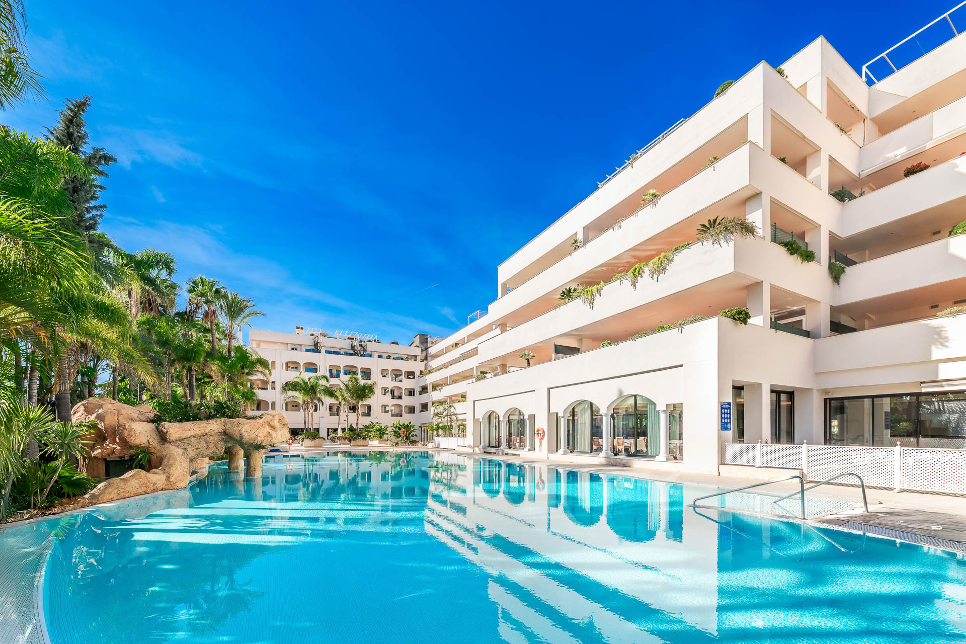 Large swimming pool with turquoise water reflecting a modern white apartment building under a clear blue sky. [AI-generated text]