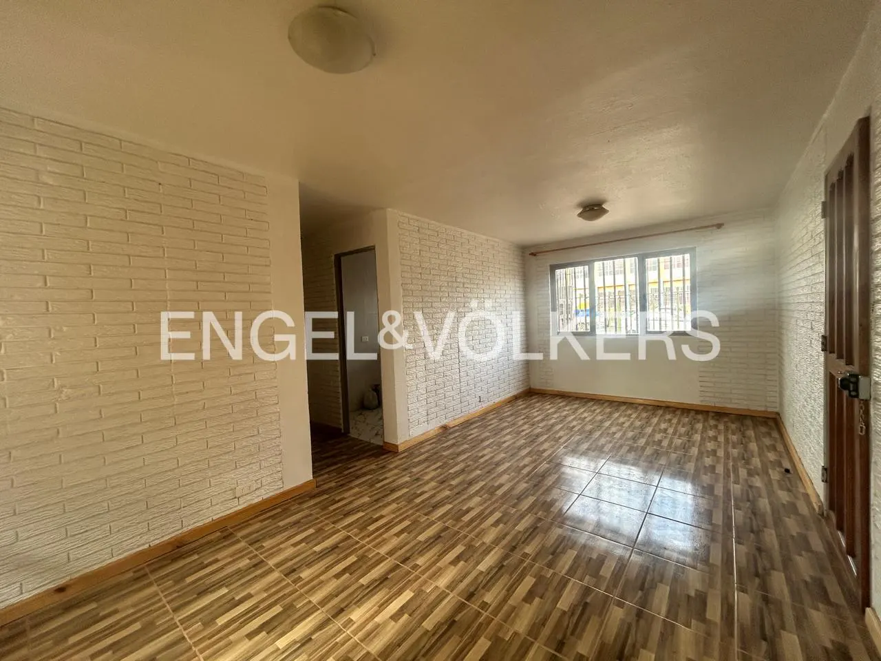 A bright, empty room with tile floors, white brick walls, and a window with bars. A doorway leads to another room.