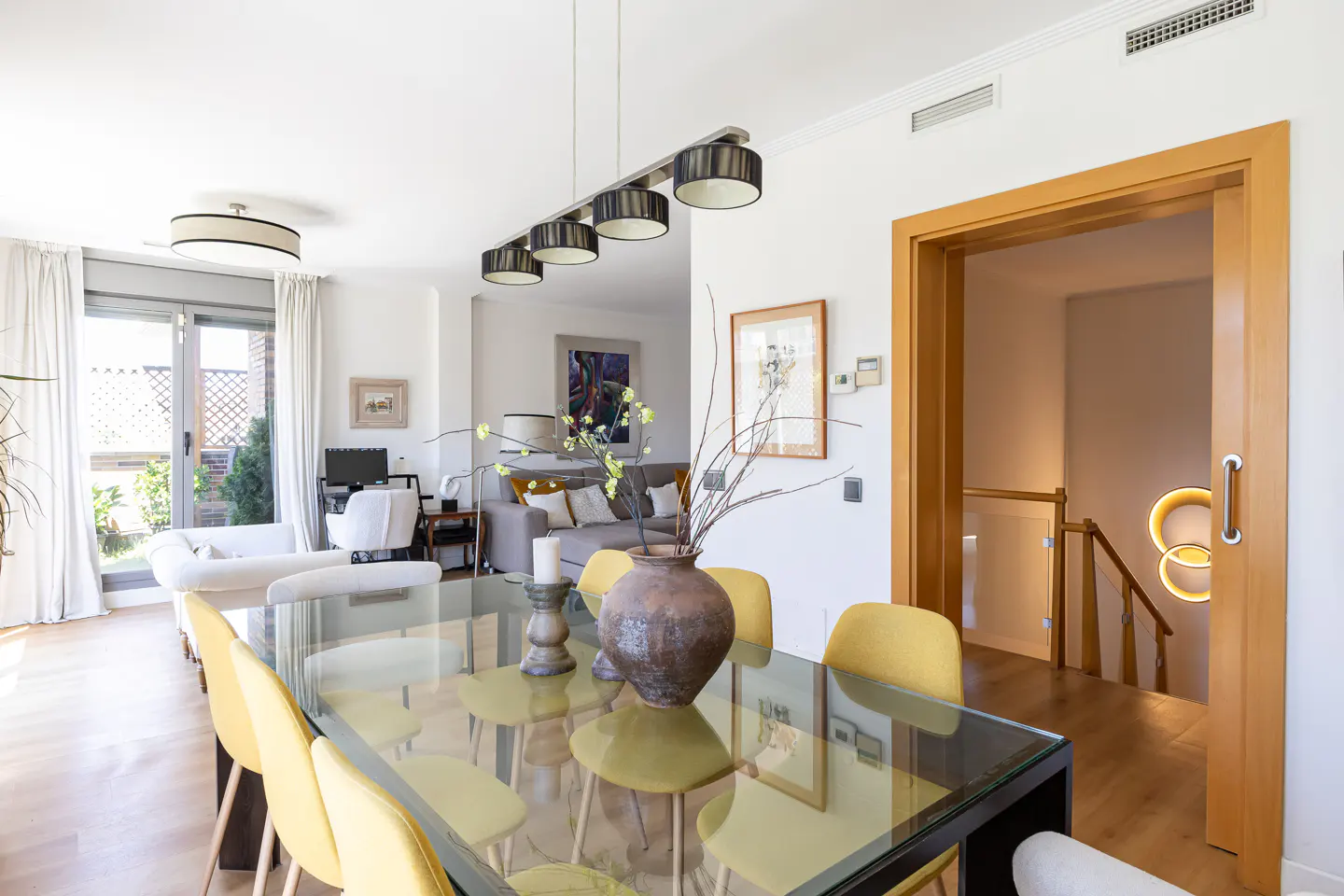Bright dining room with a glass table, yellow chairs, and a vase. A living room and staircase are visible in the background.