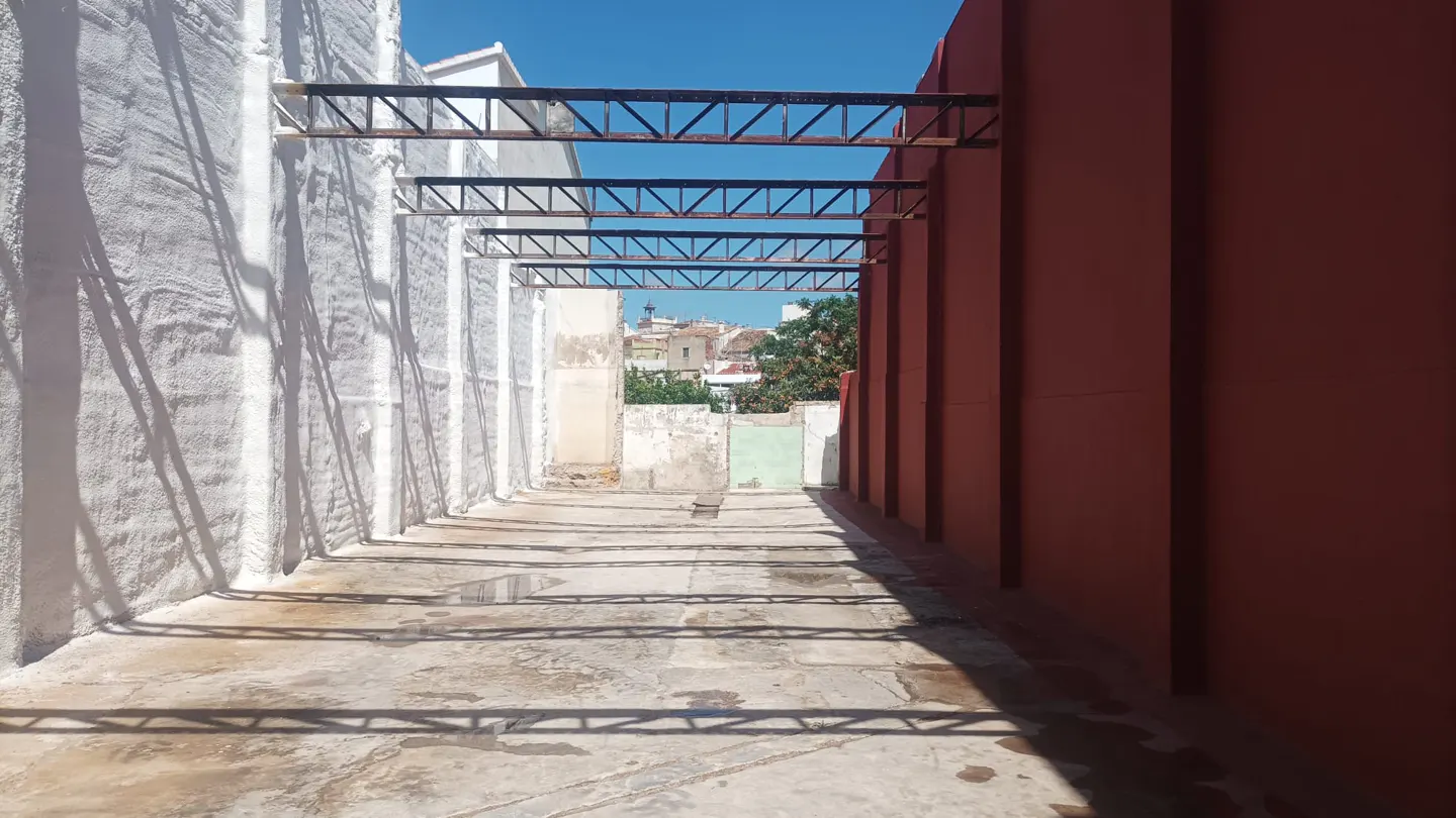 Outdoor space with metal beams casting shadows on the concrete floor, flanked by a white wall and a red wall. Cityscape visible in the distance.