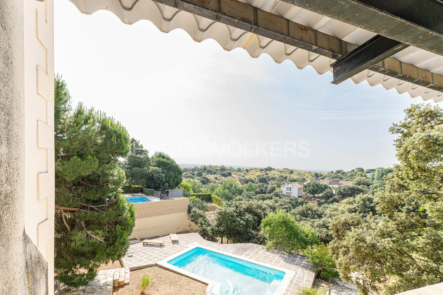 View from a balcony overlooking two blue swimming pools, green trees, and a distant house under a bright sky.