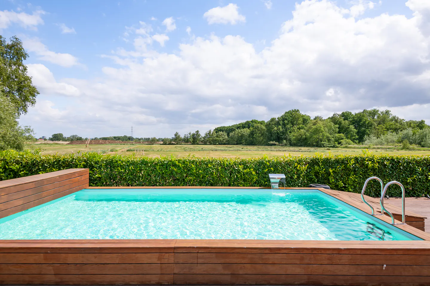 Outdoor pool with turquoise water, surrounded by wood decking and a green hedge, with a field and trees in the background.