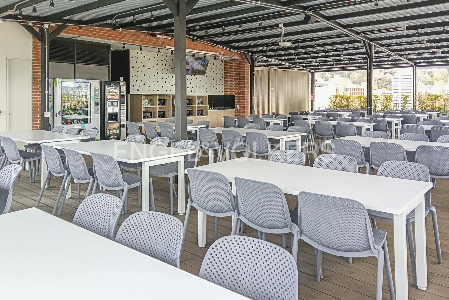 A large, open-air dining area with white tables and gray chairs under a metal roof structure. Vending machines and a brick wall are visible in the background.