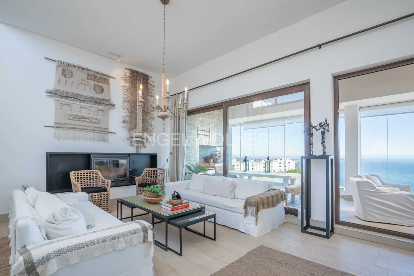 Bright living room with white sofas, woven chairs, and a fireplace. Large windows offer ocean views. A chandelier hangs above a coffee table.